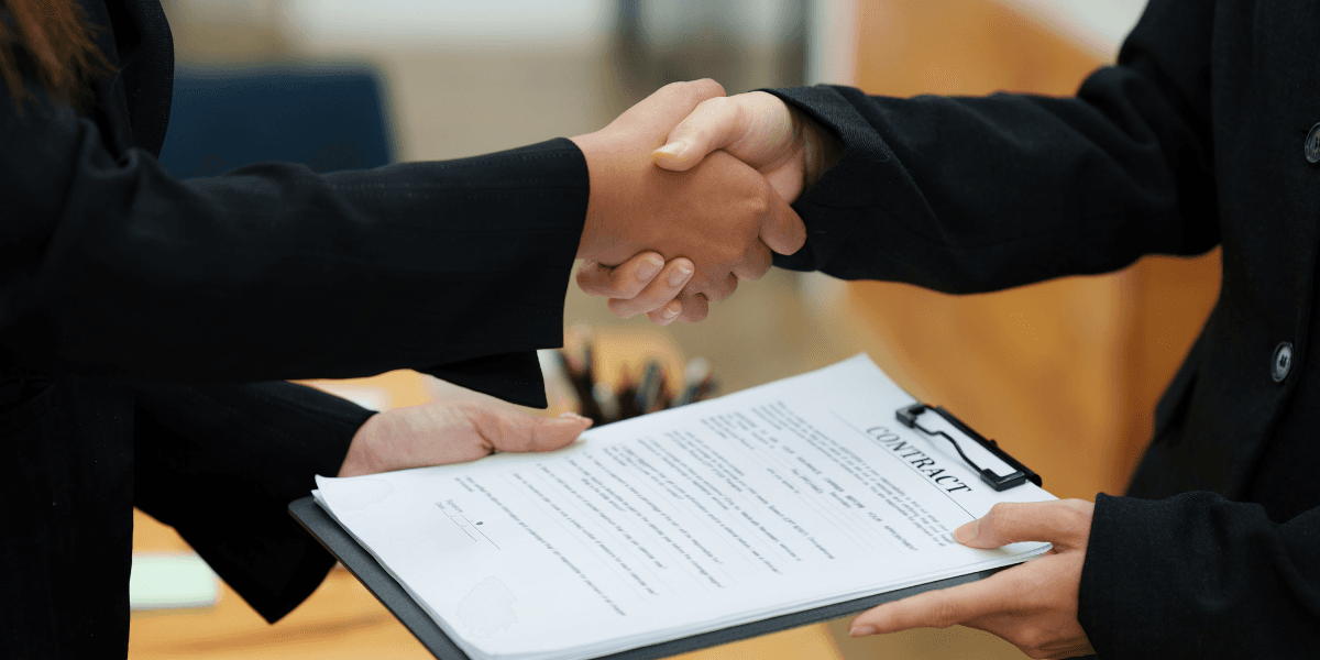Two individuals shaking hands over a contract on a clipboard in an office setting
