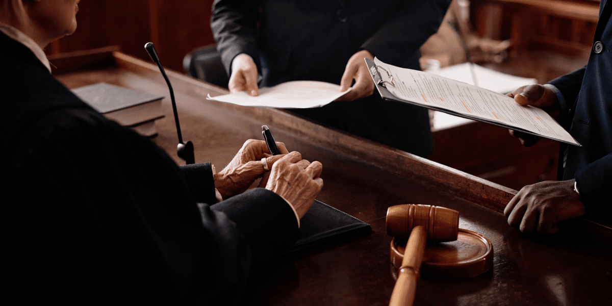 A judge writes notes while two people review documents in a courtroom setting