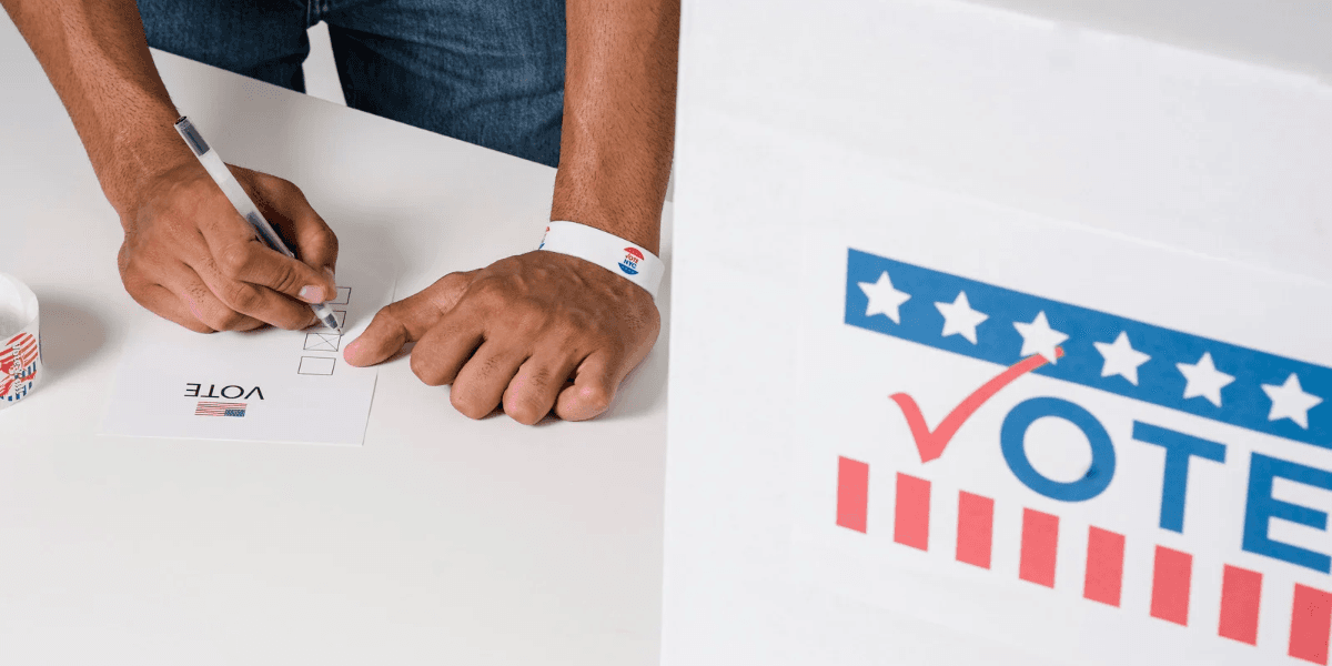 Person marking a ballot at a voting booth with a "Vote" sign visible