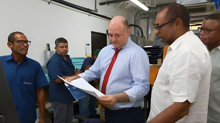 A group of five men stand in a workspace, examining a document. The man in the center, wearing a blue shirt and red tie, appears focused. The others watch attentively, suggesting a collaborative discussion. The setting is a modern office with equipment visible in the background.