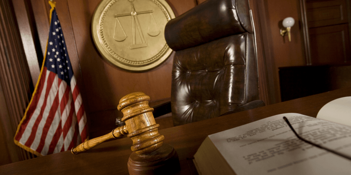 Judge's gavel on a courtroom bench with the U.S. flag and scales of justice in the background.