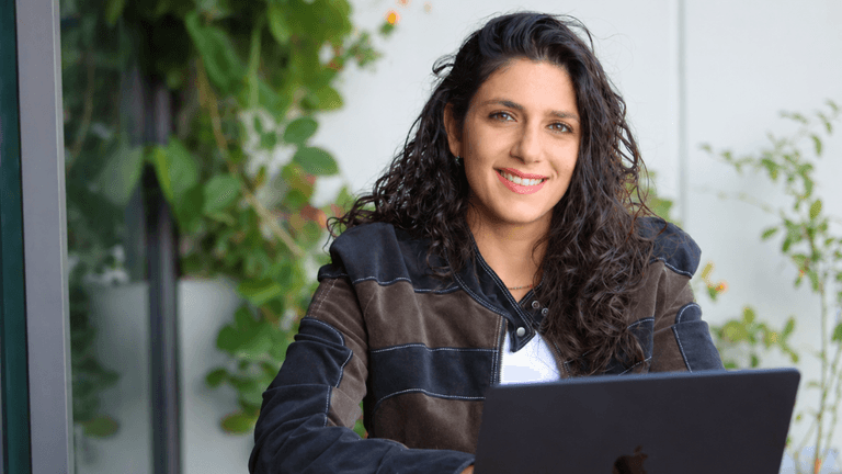 Woman smiling while working on a laptop outdoors surrounded by greenery.