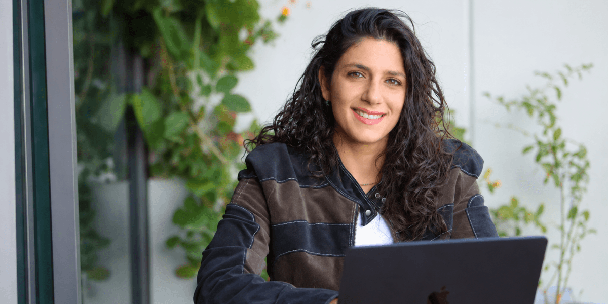 Woman smiling while working on a laptop outdoors surrounded by greenery.