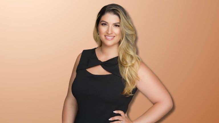 Portrait of a confident woman with long blonde hair, wearing a black dress, smiling against a warm orange background. She exudes professional charm and elegance, suitable for use in business or personal branding materials.