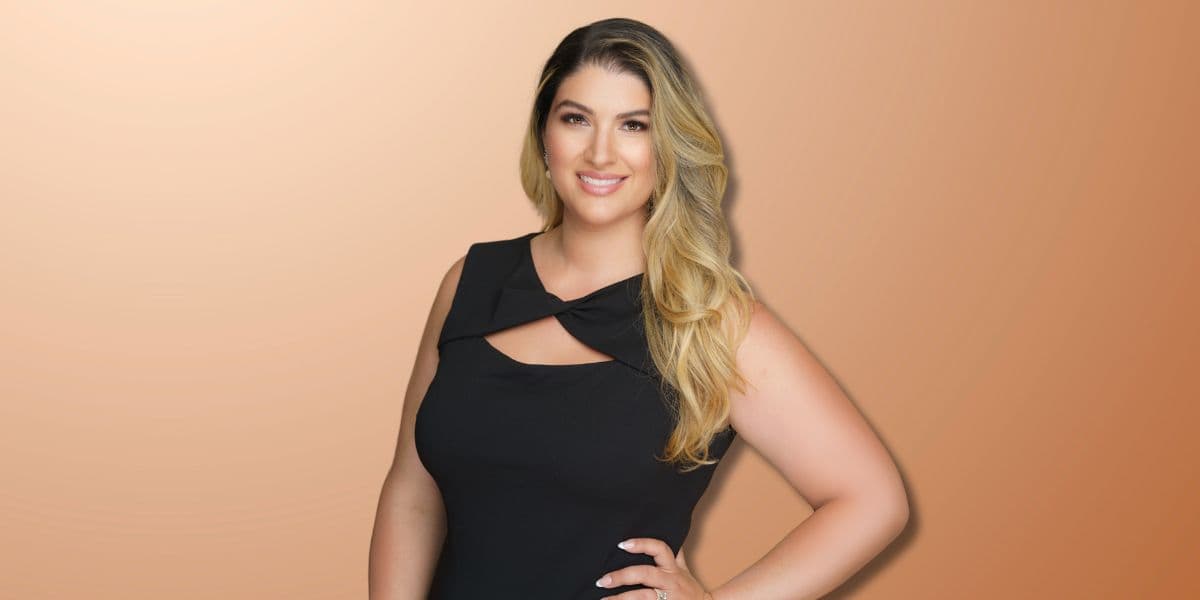 Portrait of a confident woman with long blonde hair, wearing a black dress, smiling against a warm orange background. She exudes professional charm and elegance, suitable for use in business or personal branding materials.