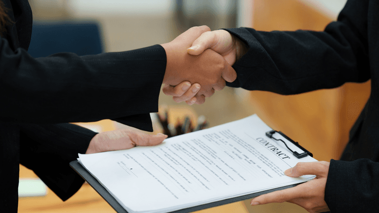 Two individuals shaking hands over a contract on a clipboard in an office setting