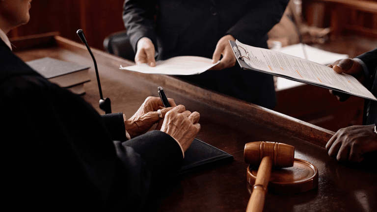 A judge writes notes while two people review documents in a courtroom setting