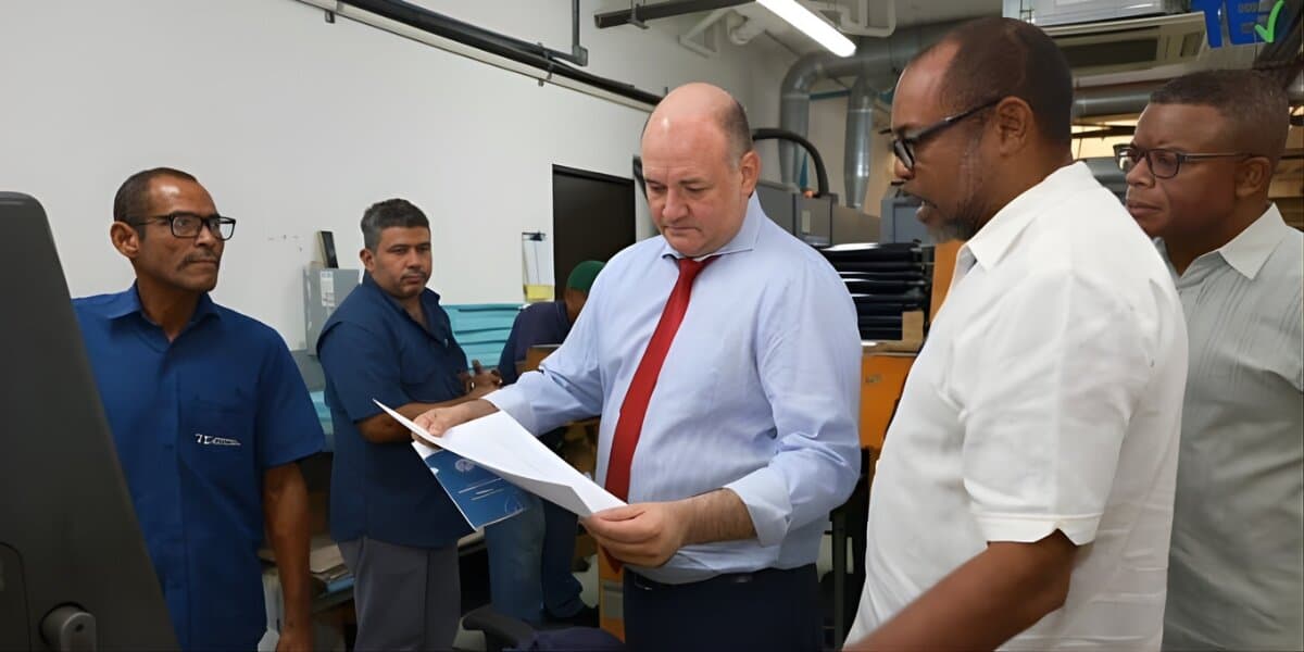 A group of five men stand in a workspace, examining a document. The man in the center, wearing a blue shirt and red tie, appears focused. The others watch attentively, suggesting a collaborative discussion. The setting is a modern office with equipment visible in the background.