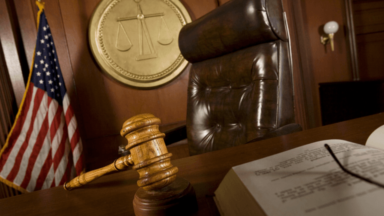 Judge's gavel on a courtroom bench with the U.S. flag and scales of justice in the background.