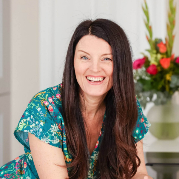 Smiling woman with long dark hair wearing a floral dress, leaning forward indoors with flowers in the background.